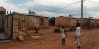Das Bild aus Südafrika zeigt RDP houses in Lehae, Johannesburg. Im Hintergrund ist ein 1 Etagen Gebäude zu sehen und im Vordergrund stehen zwei Kinder