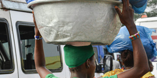A woman carries a large metal basin filled with blue bags on her head while holding a child secured on her back with a patterned cloth. She is walking along a busy street with vehicles and other people in the background.