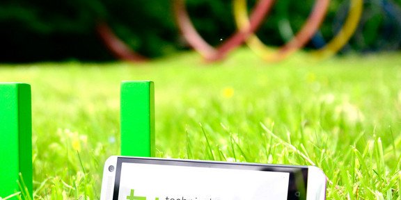 The green logo of TU Dortmund University is standing on a meadow. A smartphone is leaning against it.