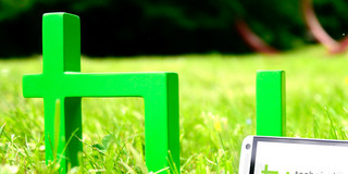 The green logo of TU Dortmund University is standing on a meadow. A smartphone is leaning against it.